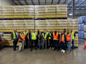 A group of logistics professionals in high-visibility safety vests standing together for a team photo in front of large, floor-to-ceiling stacks of yellow Pacifico beer cases in a Chicago warehouse.