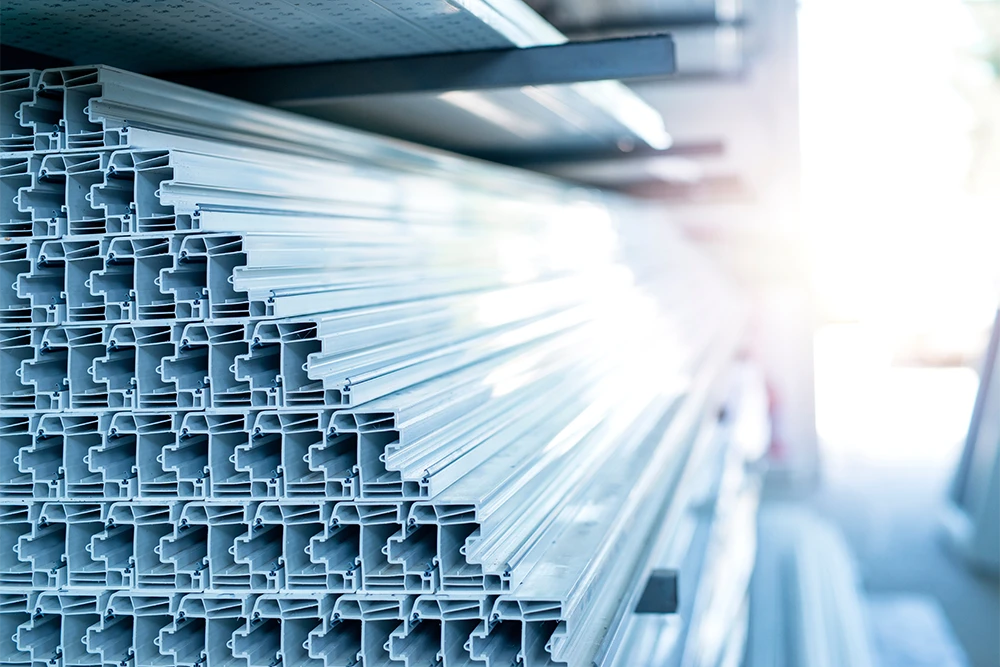 A stack of light-colored, extruded rigid PVC profiles with complex cross-sections, stored horizontally on industrial shelving in a warehouse.